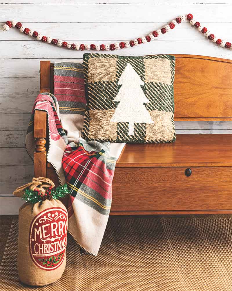 A bench with a plaid blanket, a pillow with a Christmas tree, and a pompom garland. 