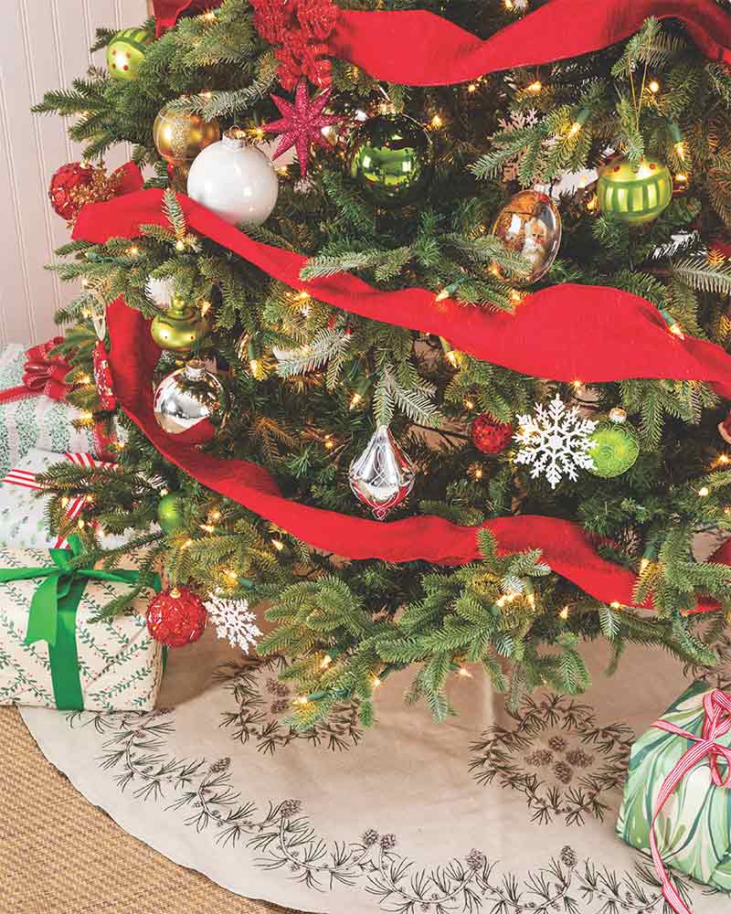 A Christmas tree with metallic ornaments and a tree skirt embroidered with pine cones. 