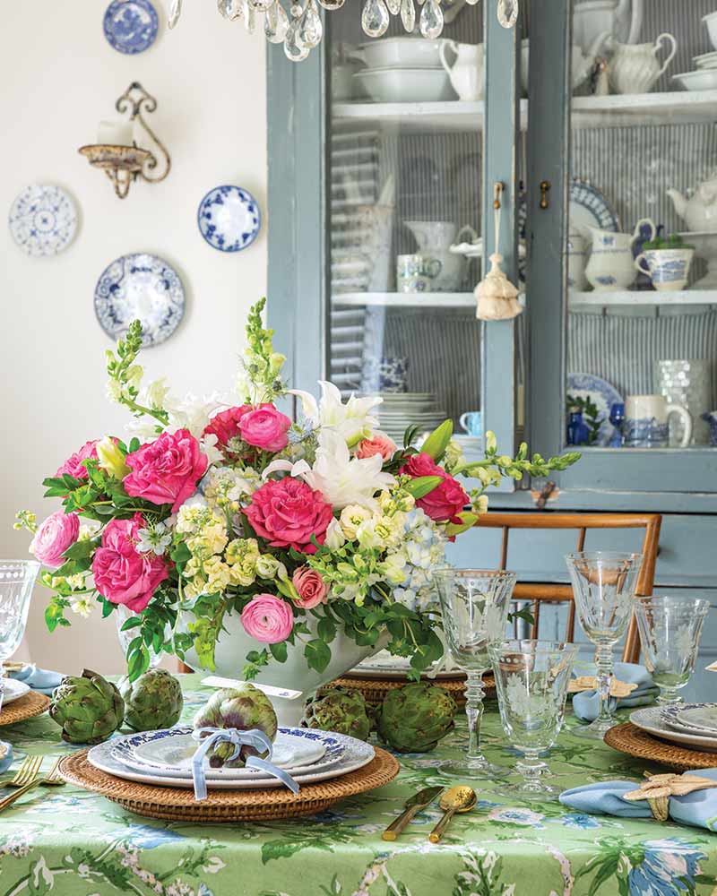 A tablesetting with a blue-and-green floral tablecloth and an arrangement of pink blooms.