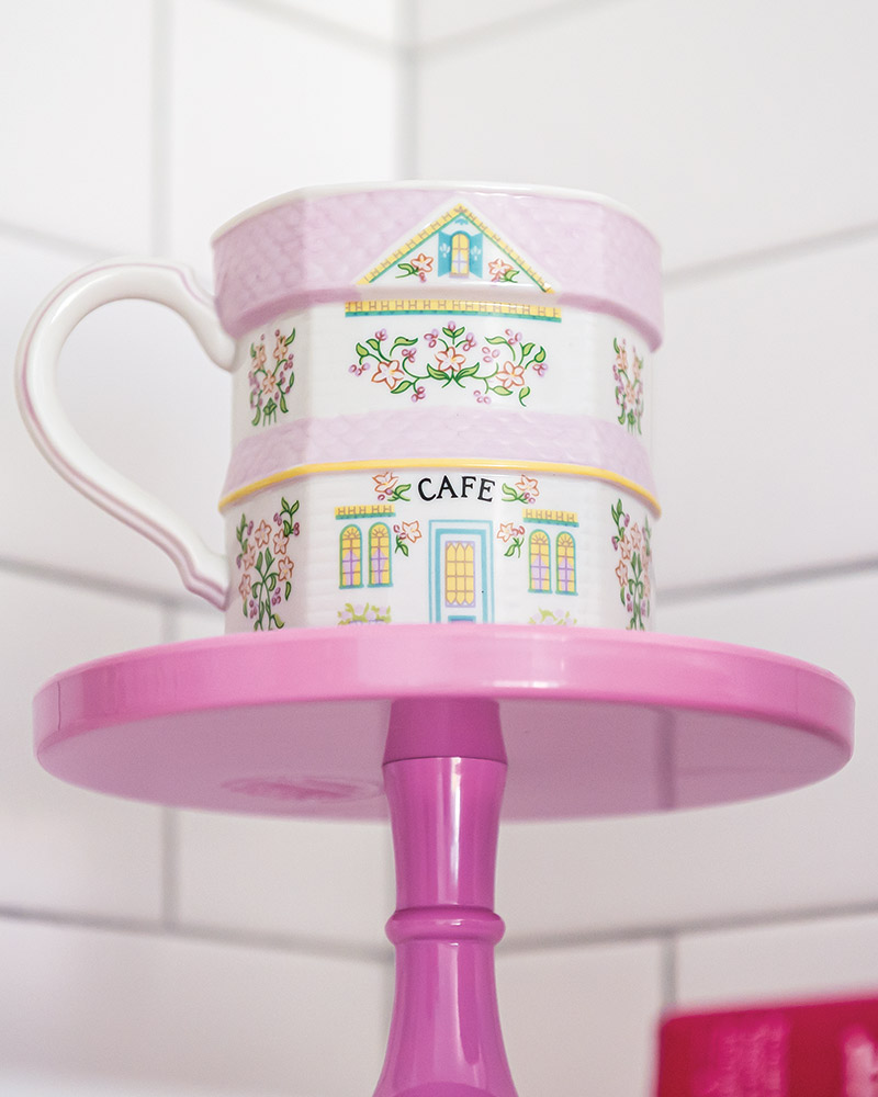 A cup from The Lenox Spice Village collection styled on a pink cakestand.