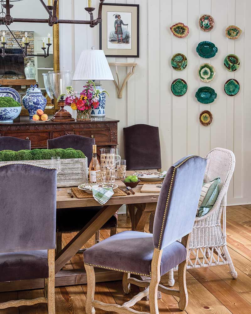 A cozy dining room featuring a rustic wooden table set with clear glasses and a colorful bouquet. Three different styles of dining chairs are placed around the table, each with varying designs and muted fabric colors. The room is decorated with a collection of green-toned decorative plates mounted on the wall and several blue and white vases placed on a sideboard. A framed picture hangs above the sideboard complementing the room’s charming aesthetic.