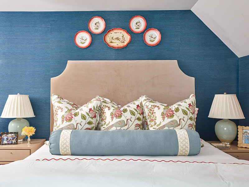 This image features an elegantly decorated bedroom with a large bed covered in floral pillows, flanked by two table lamps, against a vibrant blue wall adorned with three decorative plates.