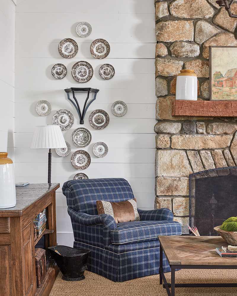 A cozy room featuring a stone fireplace on the right with a rustic wooden mantel displaying a white vase and books. A plaid upholstered armchair with a wooden horse sculpture is placed in front of a wall decorated with a symmetrical arrangement of decorative plates. To the left, a wooden sideboard holds a lamp and white pottery.