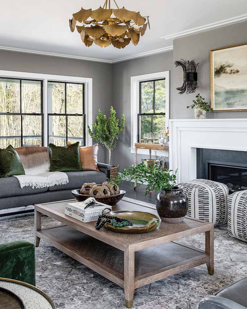 A cozy, modern living room with elegant gray walls and large windows overlooking nature, featuring a stylish chandelier, a fireplace, and tastefully arranged furniture, including a sofa, armchairs, and a wooden coffee table.