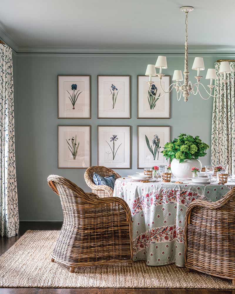 A cozy dining room featuring a round table covered with a floral tablecloth and set with dishes and a plant centerpiece. Rattan chairs face towards the table. The wall is decorated with framed botanical prints, and a chandelier hangs above. Soft, green walls and patterned curtains complete the serene ambiance.