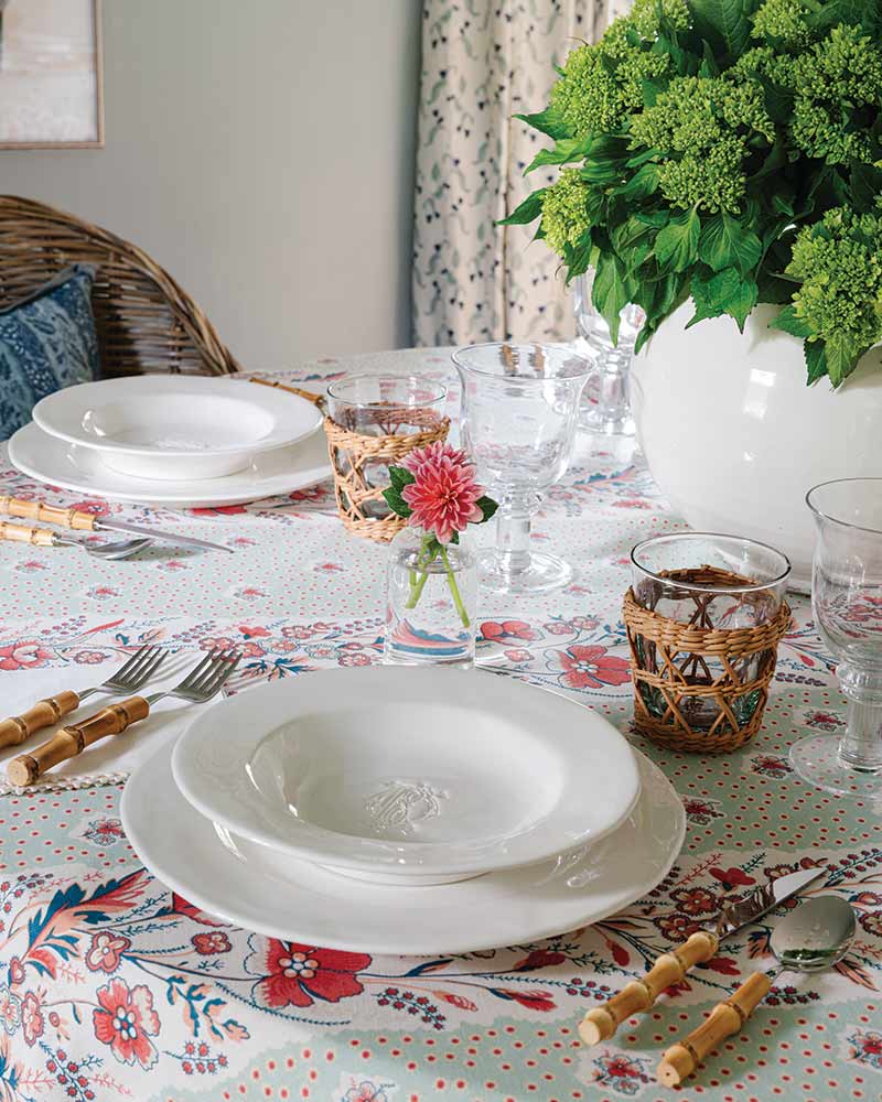 A neatly arranged dining table with floral tablecloth, featuring white plates, rattan-wrapped glasses, and a centerpiece with green hydrangeas.