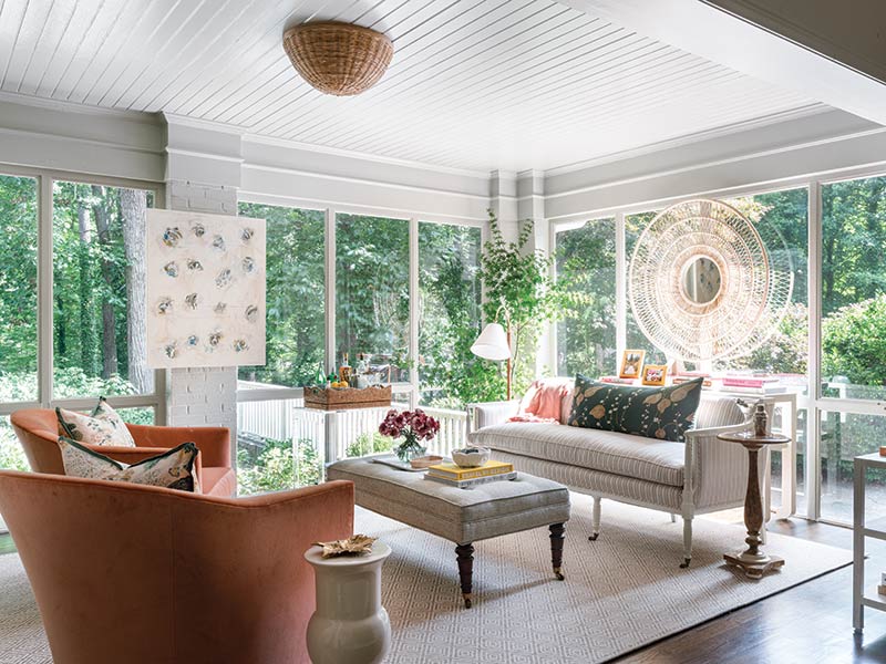 A cozy sunroom furnished with a peach armchair, a light gray sofa with assorted throw pillows, and a white coffee table. Large windows with white frames offer a view of lush greenery outside. 