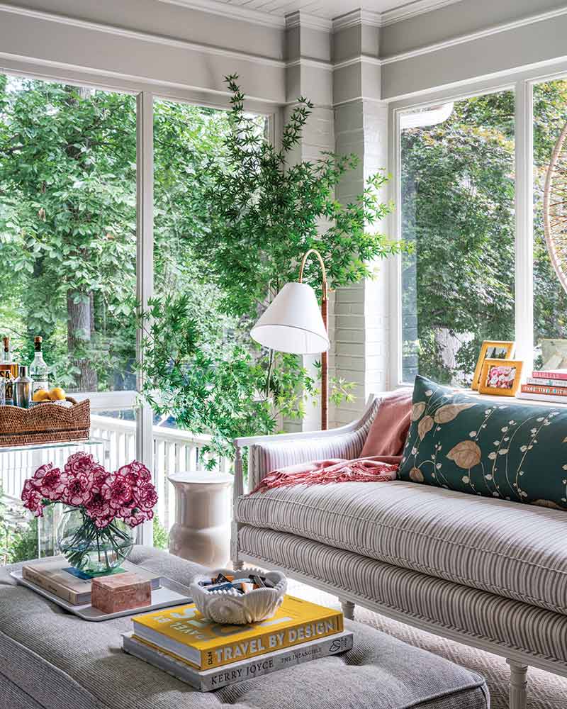 Sun porch tastefully decorated with a striped sofa adorned with various cushions, a hanging white lamp, green houseplants, and featuring a coffee table with books, including "Travel by Design" by Kerry Joyce, overlooking lush greenery visible through large windows.