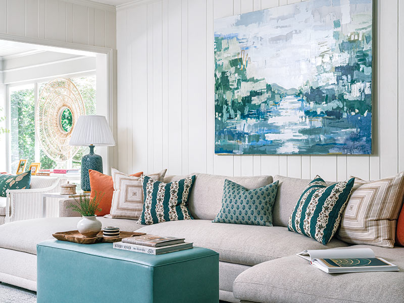 Interior of a modern living room featuring a large sectional sofa with a mix of vibrant and patterned cushions. There is a prominent blue abstract landscape painting on the wall.
