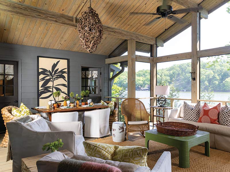 A cozy and stylish lakeside sunroom featuring large screen windows with a view of a serene lake. The room includes a variety of seating arrangements like sofas and chairs, accented with colorful pillows. A well-decorated dining table is set near a wall adorned with large botanical prints. The ceiling is wooden with visible beams and equipped with ceiling fans. The overall atmosphere is warm and inviting, blending indoor comfort with outdoor nature seamlessly.