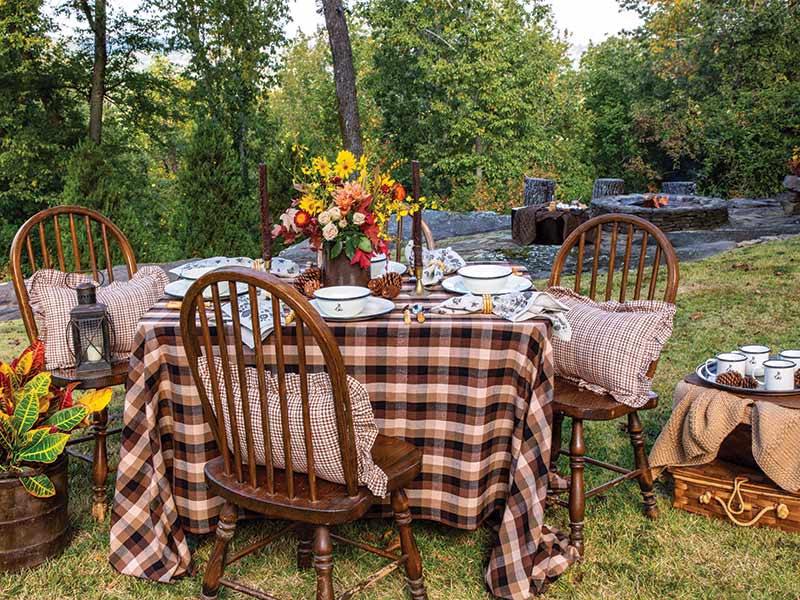 Outdoor dining setup featuring an elegantly arranged table with plaid tablecloth, fresh flowers, fine china, and wooden chairs, set in a serene garden with trees and a stone firepit.