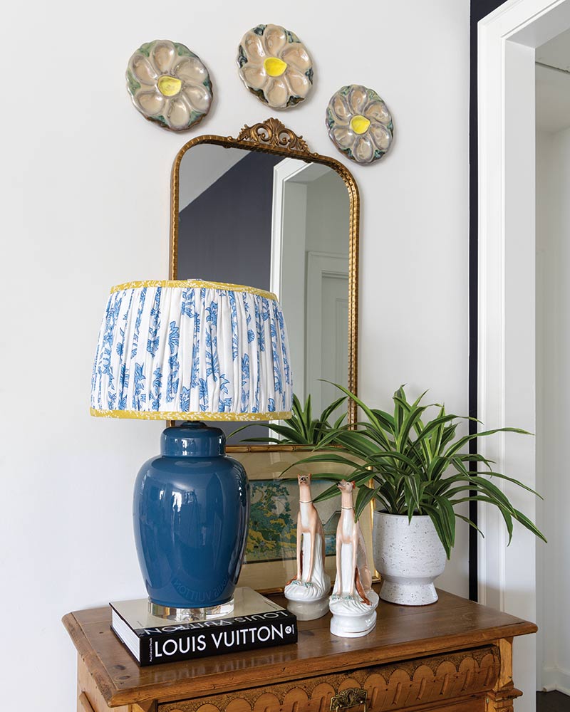 An elegant interior decor arrangement featuring a vintage-style lamp with a blue base and a shade patterned in blue and white, placed on top of a stack of books labeled "LOUIS VUITTON." Beside the lamp, there's a potted green plant. The lamp stands on an ornate wooden table, with a decorative mirror above it framed in gold. On the wall, three oyster plates are mounted above the mirror.