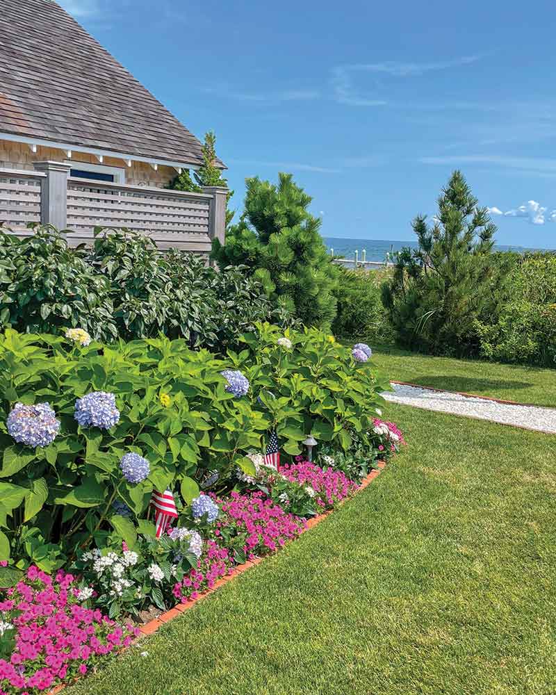 A serene garden with a view of the ocean. The garden features lush greenery, hydrangeas, and vibrant pink flowers along a neatly trimmed lawn.  Small American flags are placed intermittently among the flowers.
