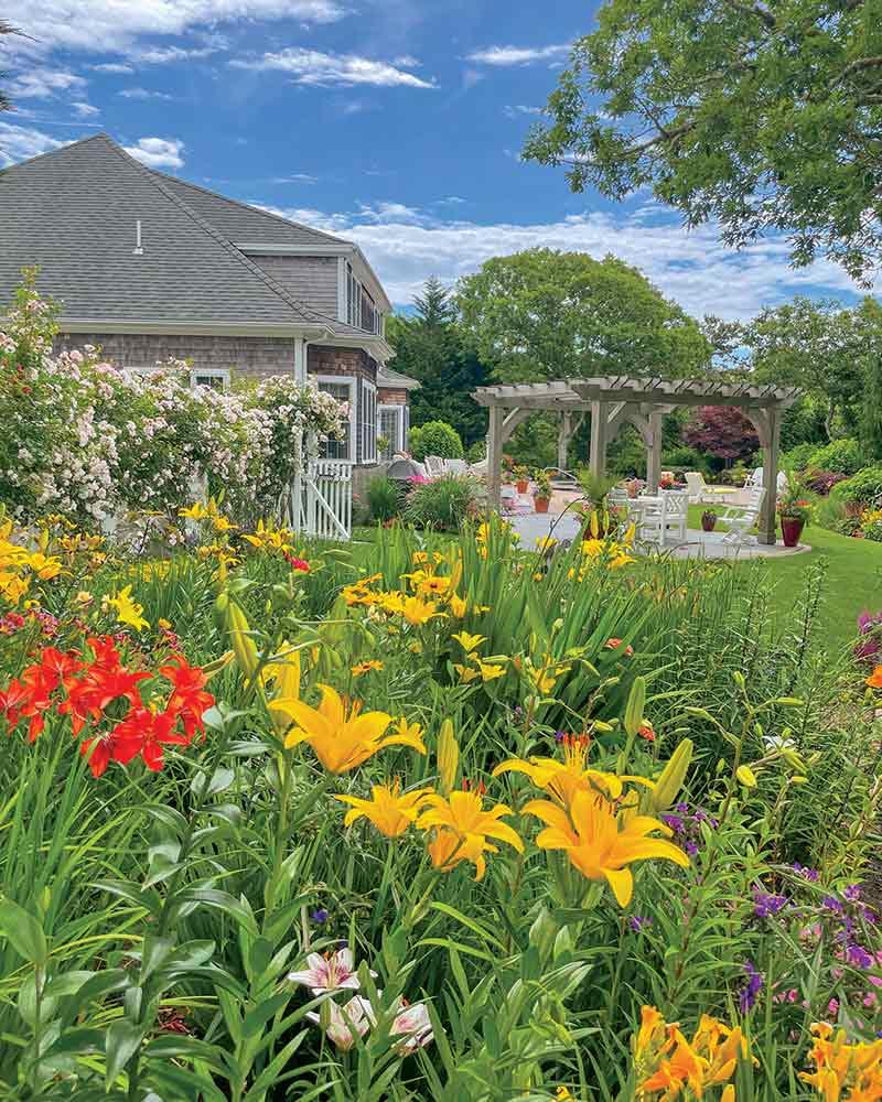A vibrant garden in full bloom with a variety of colorful flowers, complemented by a well-maintained lawn and a rustic pergola near a residential house, showcasing a serene outdoor setting.