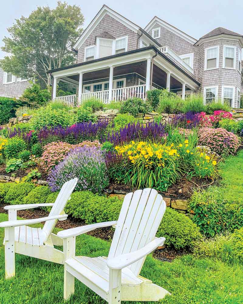 Two white Adirondack chairs facing a lush, colorful garden in full bloom in front of a traditional wooden house with a wrap-around porch.