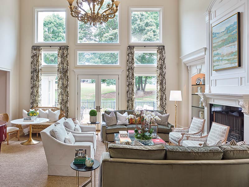 Elegant living room decorated in a classic style with a large chandelier, white sofas, and a fireplace, featuring large windows looking out to a green lawn, enhanced by tasteful landscape paintings and floral curtains.