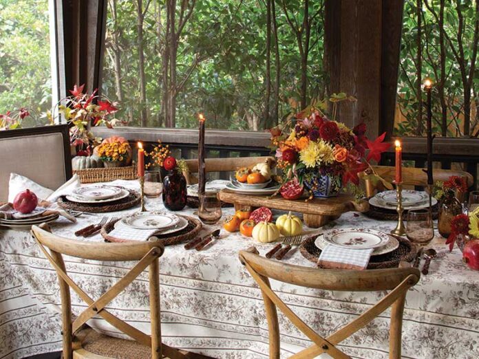 A Thanksgiving-themed dining setup with a table adorned with floral centerpieces, candles, and a fall-inspired decor, set against a backdrop of large windows overlooking green foliage.