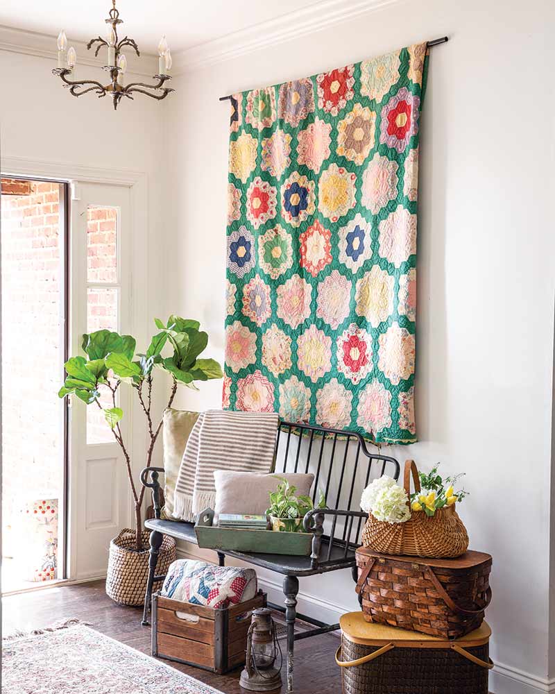 A entryway with a black bench topped with pillows and throws. A colorful vintage quilt hangs on the wall over the bench.
