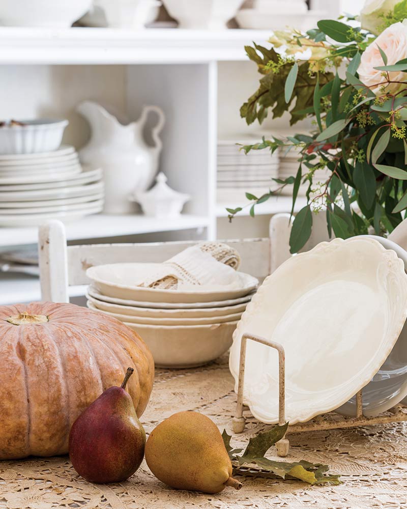 A tabletop with ironstone pieces stacked beside pumpkins and pears.
