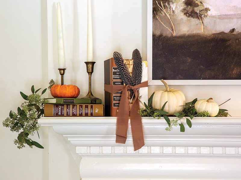 Decorative mantelpiece with an arrangement of elements including books, a small painting of a landscape, two white candles, a brass candlestick, a pair of feathers tied with a brown ribbon, and several pumpkins surrounded by green foliage.