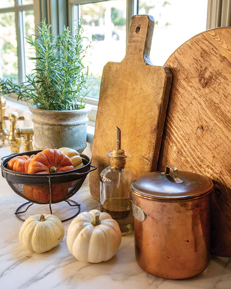 A kitchen setting featuring rustic wooden cutting boards, a copper container, an oil dispenser, and a metal basket with pumpkins. Decorative elements include white miniature pumpkins and a potted plant on a sunlit countertop.