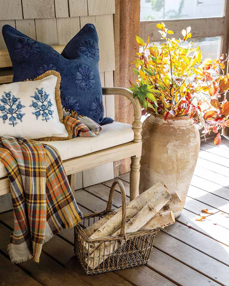 A cozy porch setting with a white wooden chair adorned with floral patterned blue and white pillows, next to a large terracotta vase holding bright yellow and red foliage. A plaid blanket drapes over the chair arm, and a wicker basket with birch logs rests nearby.
