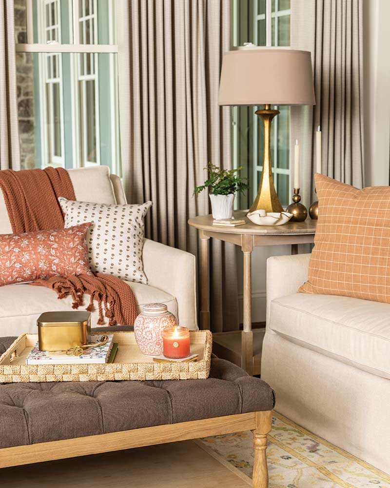 A cozy living room setting featuring an elegantly arranged sofa with various patterned pillows, a stylish coffee table with decor items including candles and books, complemented by a standing lamp and striped curtains in the background.