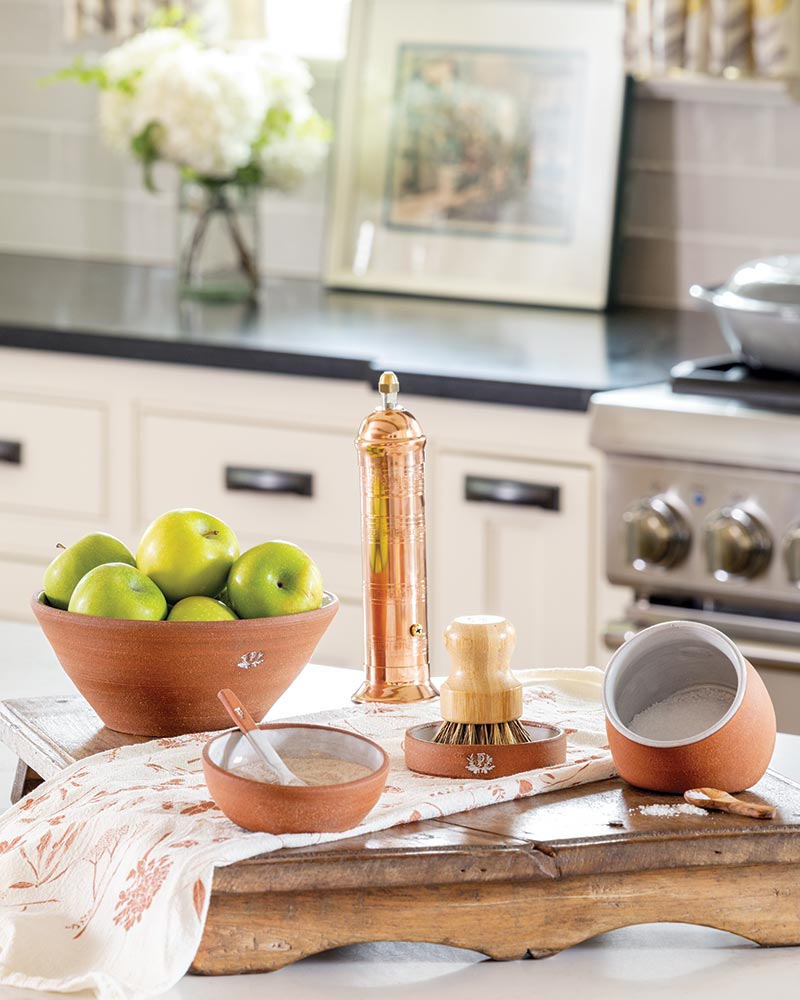 A kitchen countertop is adorned with various items that create a charming domestic scene. In the foreground, a wooden tray holds a bowl filled with green apples, next to a small bowl containing salt and a textile beneath them features a floral pattern. A copper pepper mill stands tall behind the fruit bowl. In the background, part of a white kitchen cabinet is visible, and a framed picture rests against a tiled backsplash. The setting exudes a warm, inviting ambiance typical of a home kitchen.