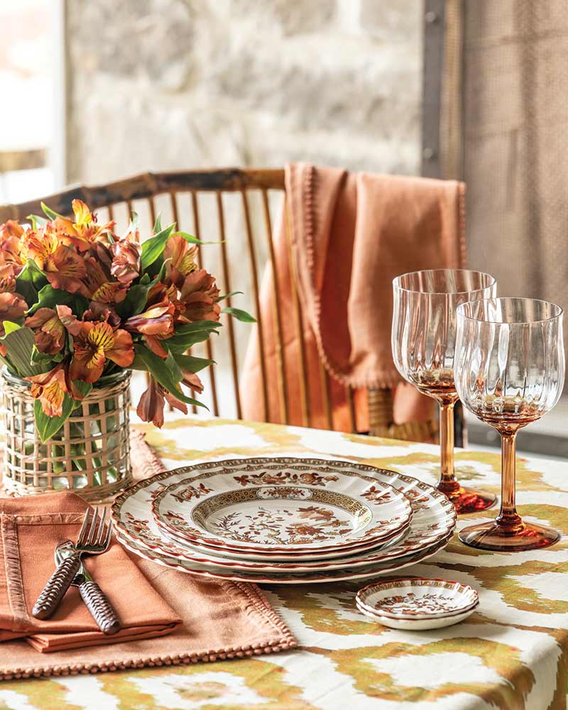 Elegantly set dining table featuring a stack of ornate, patterned plates, crystal wine glasses, and folded burnt orange napkins with copper-colored cutlery. A vase with lush, orange flowers complements the autumn-themed tablecloth. Warm, natural light enhances the inviting ambience.