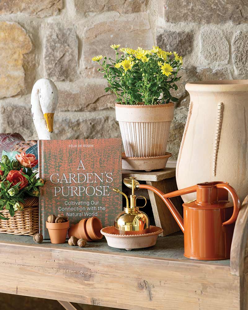 A rustic garden setting with a textured stone wall backdrop. A wooden table holds a book titled "A Garden's Purpose: Cultivating Our Connection with the Natural World" by Felix De Rosen. Beside the book, there's a vibrant yellow potted plant, a golden plant mister, an orange watering can, and a wicker basket containing various garden tools and vegetables. An elegant bird sculpture stands on one side.
