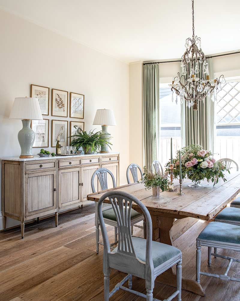 An elegantly set dining room featuring a rustic wooden table lined with gray chairs, a sideboard adorned with lamps and floral arrangements, and a chandelier, all under natural light coming in through large windows draped with green curtains.