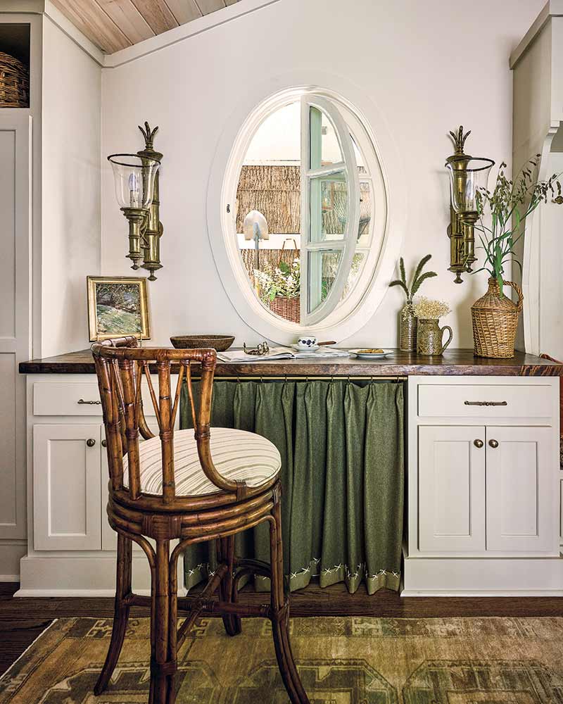 A desk under an oval window with lower cabinets and a skirted cover. A bamboo-style chair sits in front of the desk.