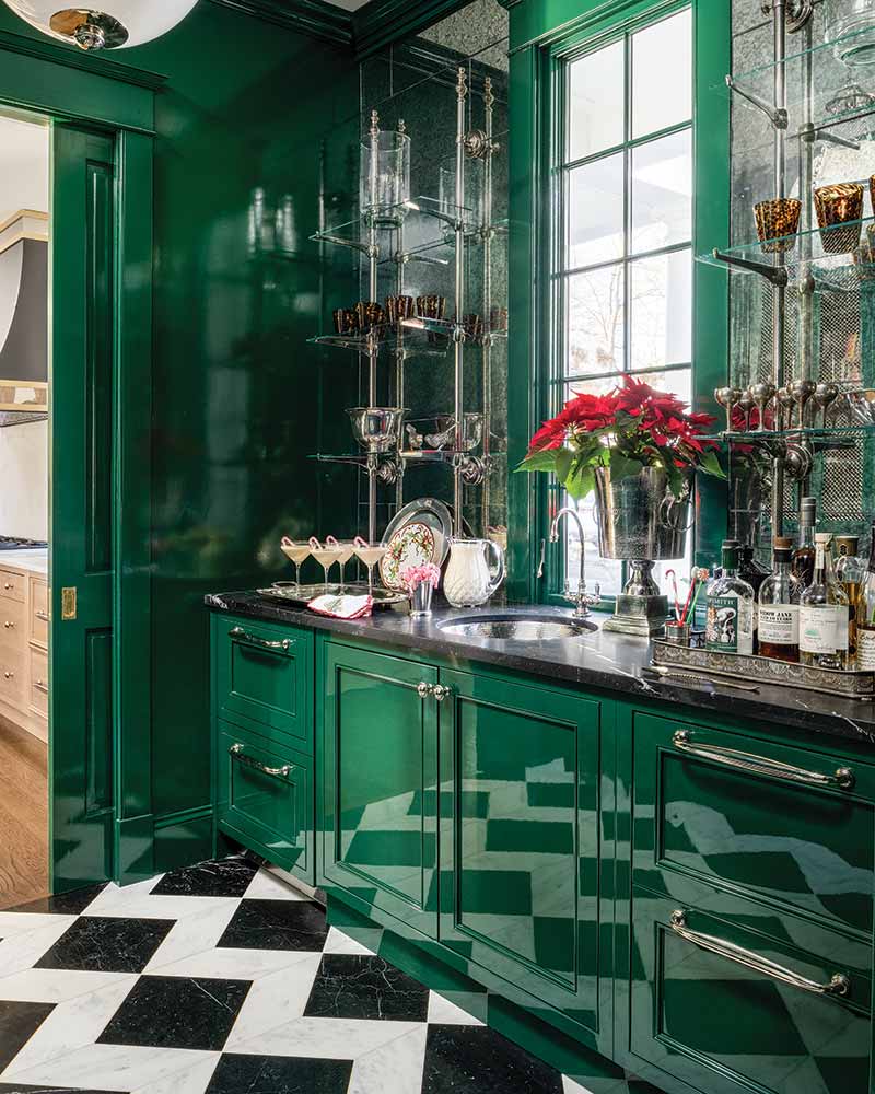 A luxurious butler's pantry featuring glossy dark green cabinetry and a black-and-white checkered floor. The room includes a built-in glass cabinet with various bottles and glassware, complemented by natural light streaming through tall windows. A poinsettia plant adds a touch of color to the space.