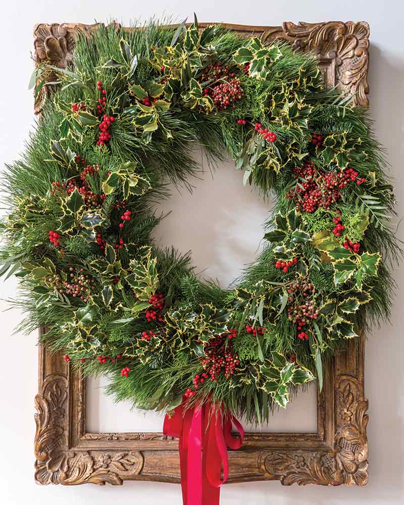 A festive Christmas wreath with green foliage and red berries, adorned with a bright red ribbon, displayed within an ornate, golden frame on a light-colored wall.