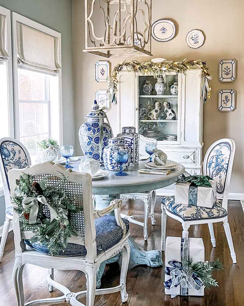 Elegant dining room decorated with a blue and white color scheme, featuring a round table set for two with vintage-style chairs, and a china cabinet adorned with festive greenery. Decor includes blue-and-white ceramic pieces and wall plates, enhancing a cozy, inviting ambiance.