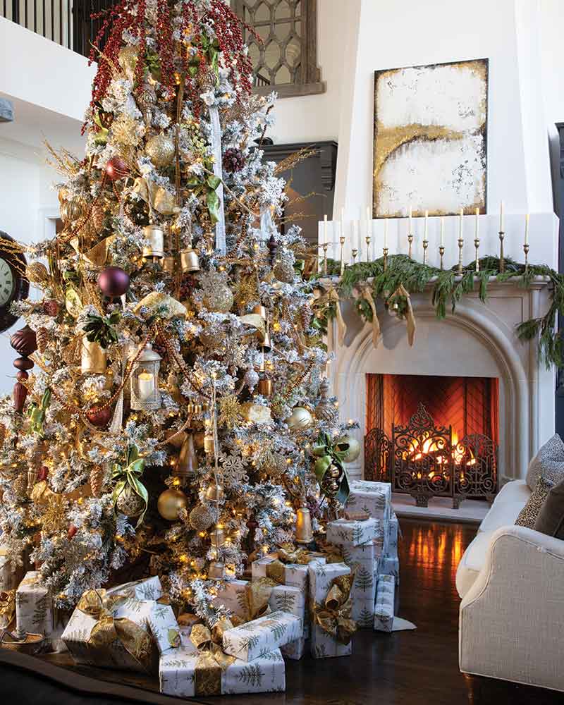 A lavishly decorated Christmas tree with gold and red ornaments and lights, set in a cozy room with a lit fireplace and elegant furnishings.