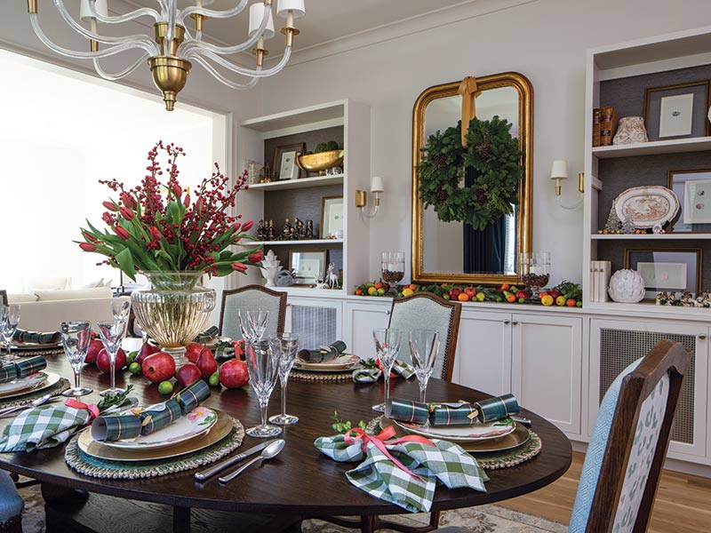 Elegant dining room decorated for a festive meal with a dark wood table set for six, featuring plaid napkins, crystal glassware, and a centerpiece of red ilex berries and pomegranates. A large gold-framed mirror and white built-in shelves filled with books and fine china add to the room’s sophisticated charm.