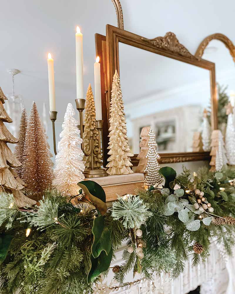 A festive holiday display featuring a variety of decorative trees of different styles and sizes on a mantelpiece, surrounded by a lush garland with green leaves and pine cones. Three taper lit candles add warmth to the scene, reflected in a large ornate mirror above the mantel.