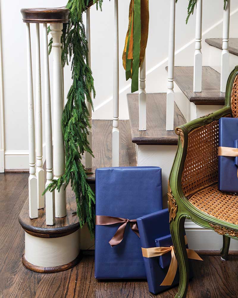 An elegant indoor scene featuring a staircase adorned with a garland, next to a vintage cane chair. Two neatly wrapped navy blue gift boxes with brown ribbons are placed on the wooden floor.