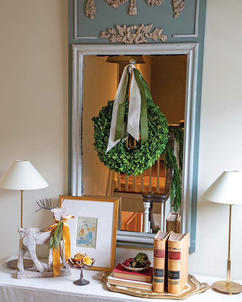 An elegantly decorated space featuring a vintage mirror with a boxwood wreath hanging on it, flanked by two lamps. In front, there is a small table displaying an assortment of items including framed artwork, an ornamental deer, and a stack of books.