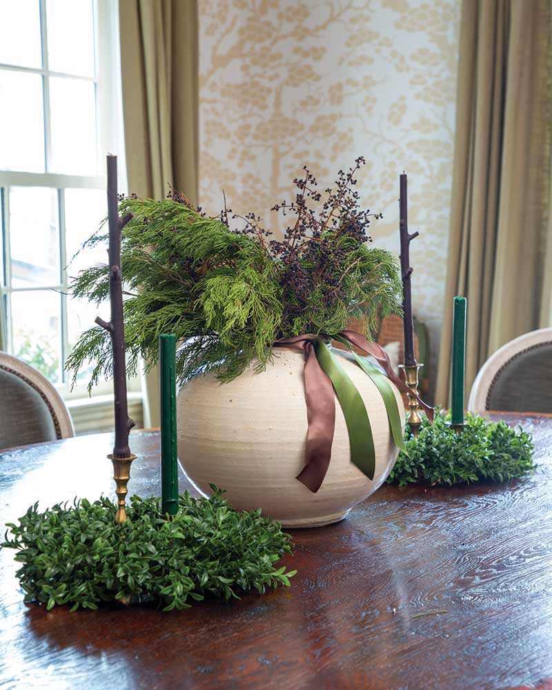A large ceramic vase with a ribbon, filled with lush greenery and branches, sits on a wooden table in an elegantly wallpapered room with large windows.
