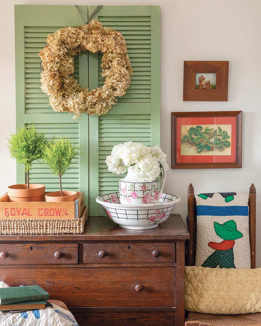 Atop the sage green shutters, a wreath composed of earthy materials draws attention, blending countryside beauty with the comforts of cottage living. A vintage-inspired feeder crate filled with vibrant potted plants shows off its lush foliage beside a vessel adorned with a delicate pink floral pattern. The pitcher, brimming with a milky white bouquet, adds tranquility to the scene, poised to leave guests with a sense of warmth and welcome.