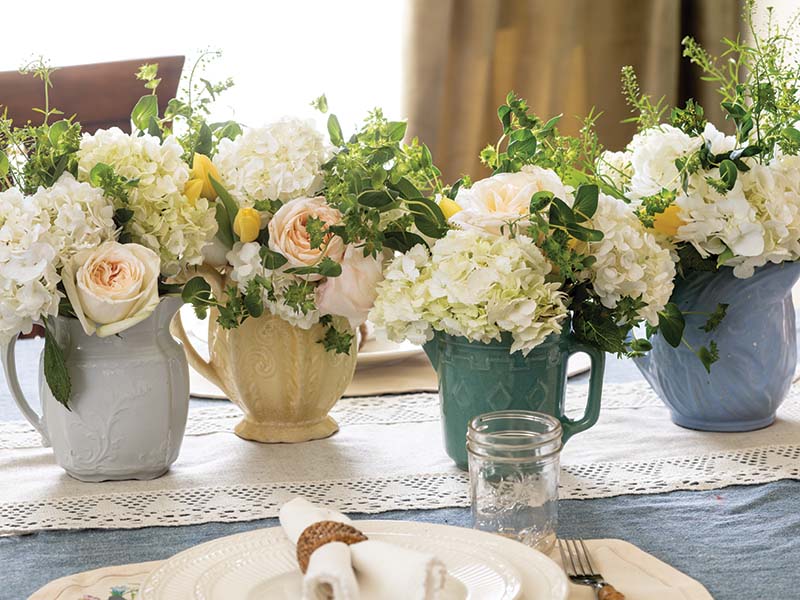 Miniature pitchers in soothing shades of baby blue, periwinkle, teal, and pale yellow line the table, overflowing with an abundance of fresh white and yellow blooms and sprigs of greenery. A lace table runner adds a touch of classic elegance reminiscent of tradition and refinement. Beneath it, a soft blue tablecloth grounds the display, harmonizing with the gentle palette above.