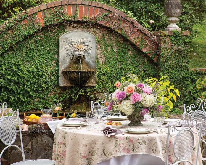 A dining table adorned with a bouquet of fresh blooms in soft shades of lilac, blush, coral, and cream beckons guests to enjoy an enchanting alfresco experience amid a backdrop of lush greenery.