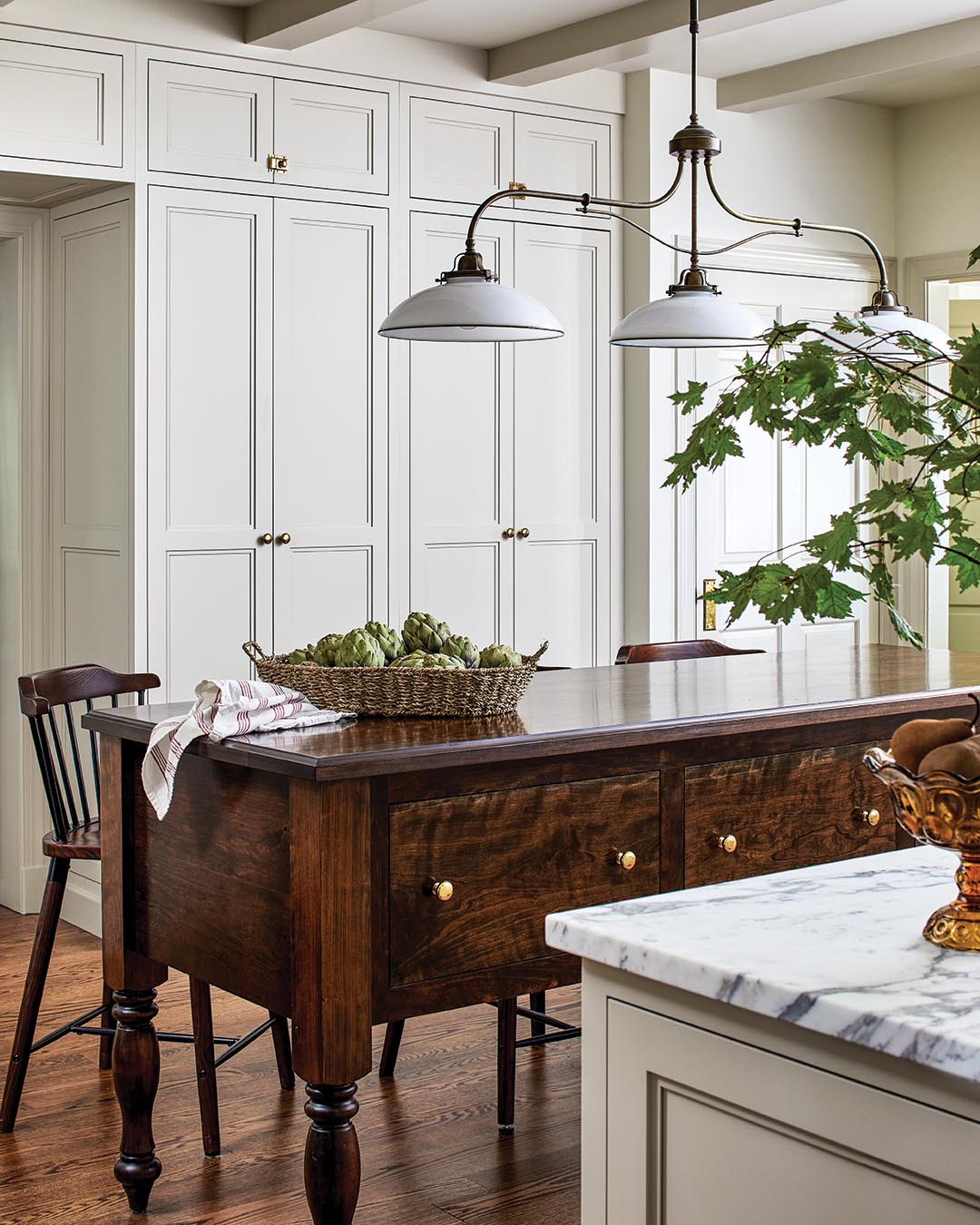 Dark wood furnishings, like the eye-catching antique island, are softened by a linen Roman shade and an Oriental rug. Streamlined cream-hued cabinetry and sleek cool-toned Arabescato Corchia marble countertops contrast with warm mixed metals throughout.