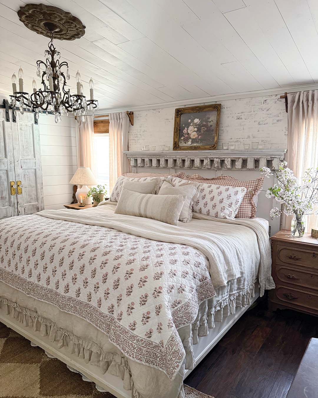 The primary bedroom boasts an accent wall with faux brick topped with a German schmear. Incorporating a salvaged piece of dentil molding, Kassie built the headboard. Gingham pillow covers pair with a quilted floral block print and ruffled linens for a layered look that offers a soothing retreat.