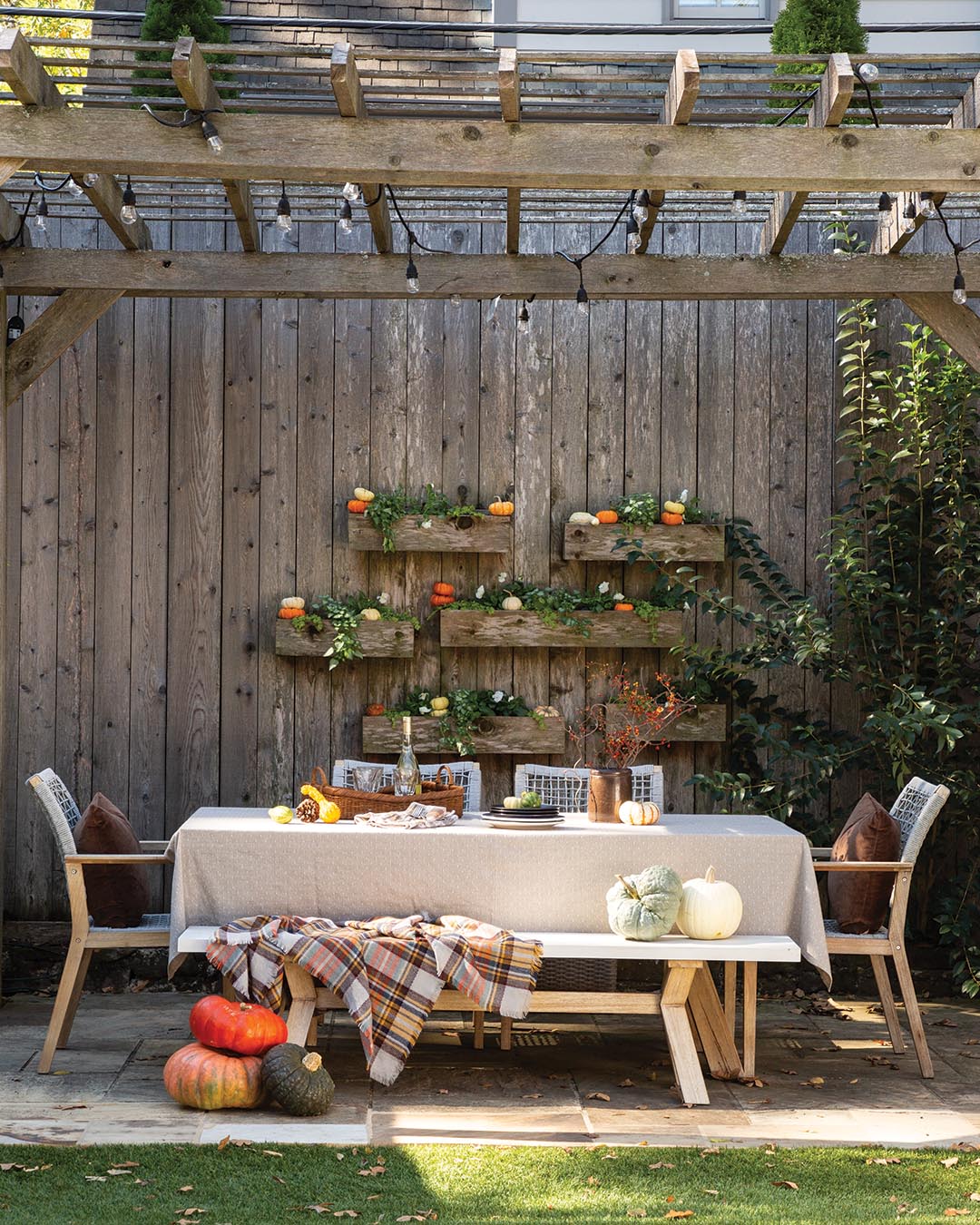 The stone patio and wooden trellis were scaled back and now play host to the outdoor dining spot. Bistro lights and a warm throw ensure the space can be enjoyed long after the sun sets. A grouping of planters built into the fence creates a captivating display for plants and seasonal accents such as miniature pumpkins.