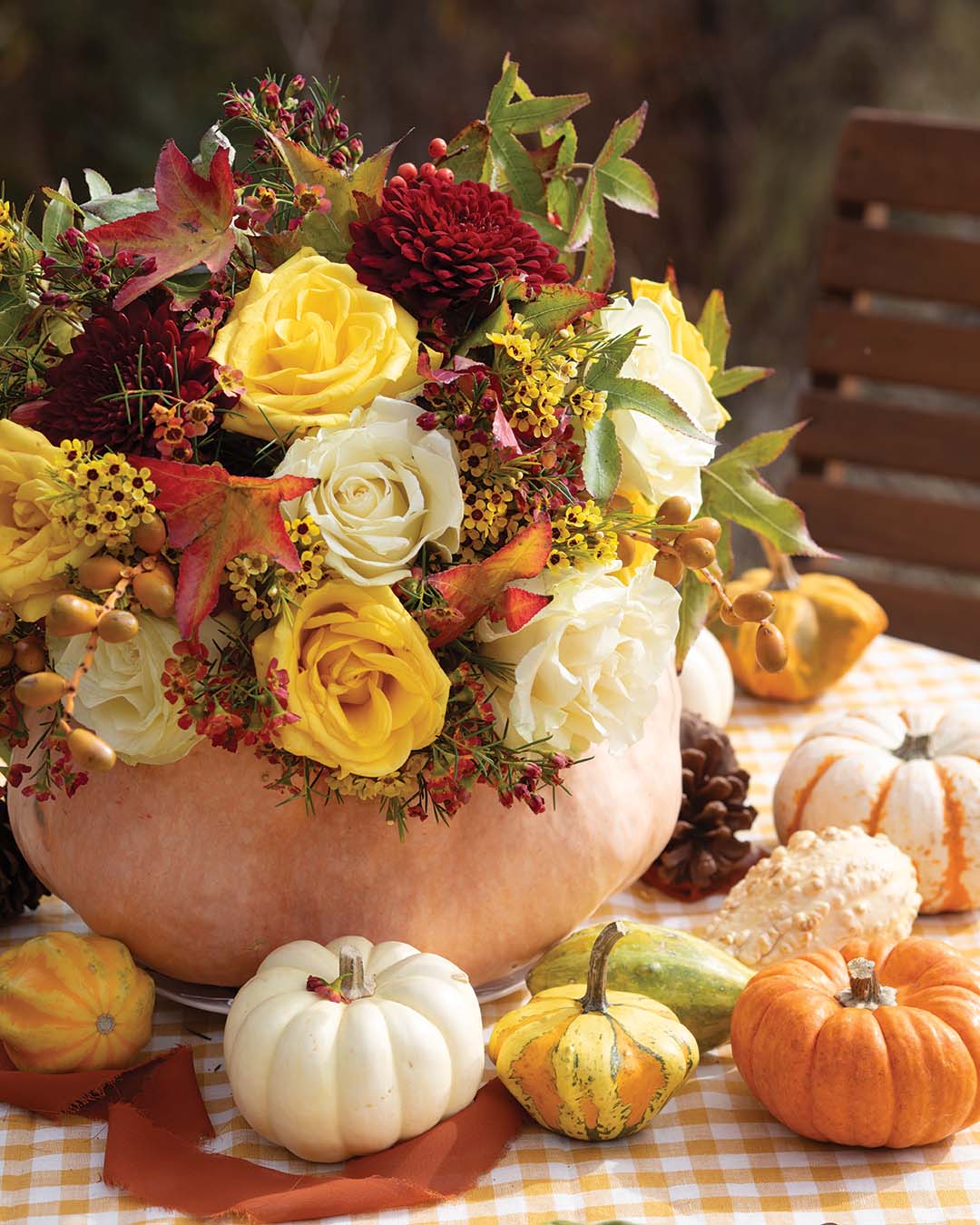 Capturing the brilliant essence of fall, a bouquet of scarlet, gold, and cream roses catches the eye, their vibrant petals standing out amidst the backdrop of lush greenery. Nestled among them is a classic token of the season, as miniature pumpkins in a spectrum of ivory, honey, and rust hues adorn the table.