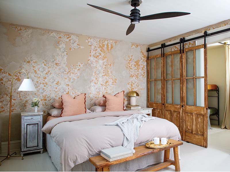 Another creative touch is the set of weathered doors mounted on tracks in the primary bedroom, likely salvaged from a school or library. The doors separate the main bedroom from a pass-through space that houses bunk beds, offering additional sleeping spaces while preserving guests’ privacy. Backed by the same wallpaper that graces the living area, the bed is piled with linen bedding that’s especially welcoming in the glow of natural light that reflects off the painted floors.