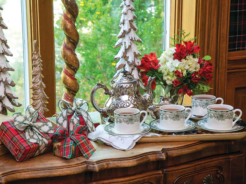 A glimmering silver teapot, accompanied by matching cream and sugar dishes, sits atop a wooden sideboard. Teacups and saucers graced with a festive poinsettia and holly design complete the tableau, ready for guests to finish the evening with a cup of tea by the fire.
